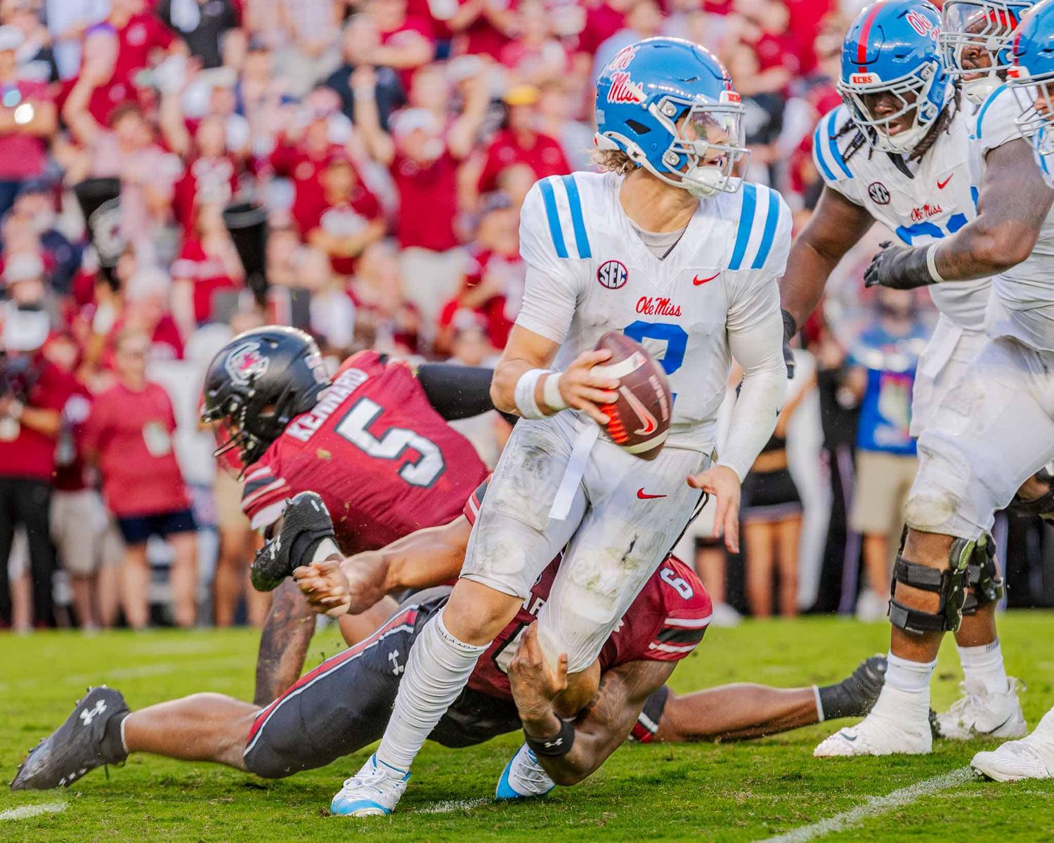 An Ole Miss quarterback in a white and blue jersey looks to pass while being pursued by a defender.