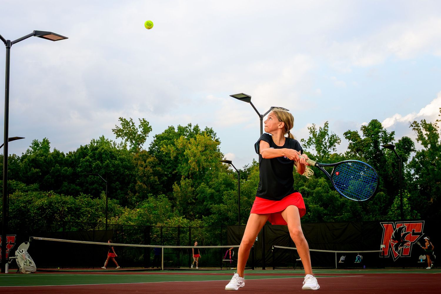 A young female tennis player in a red skirt preparing for a backhand return on an outdoor court.