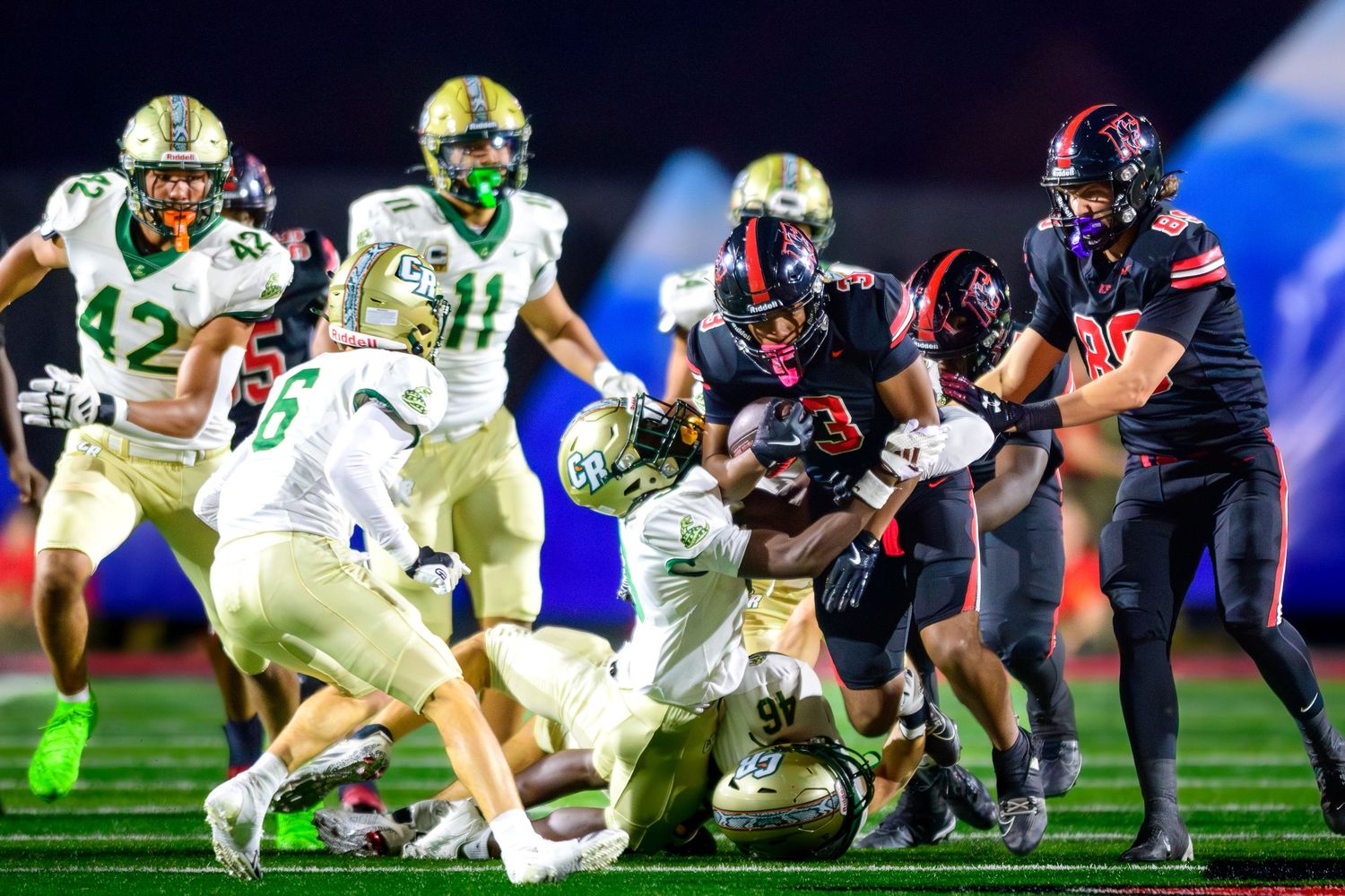 Football player in a black and red jersey being tackled by multiple defenders in gold helmets and white jerseys.