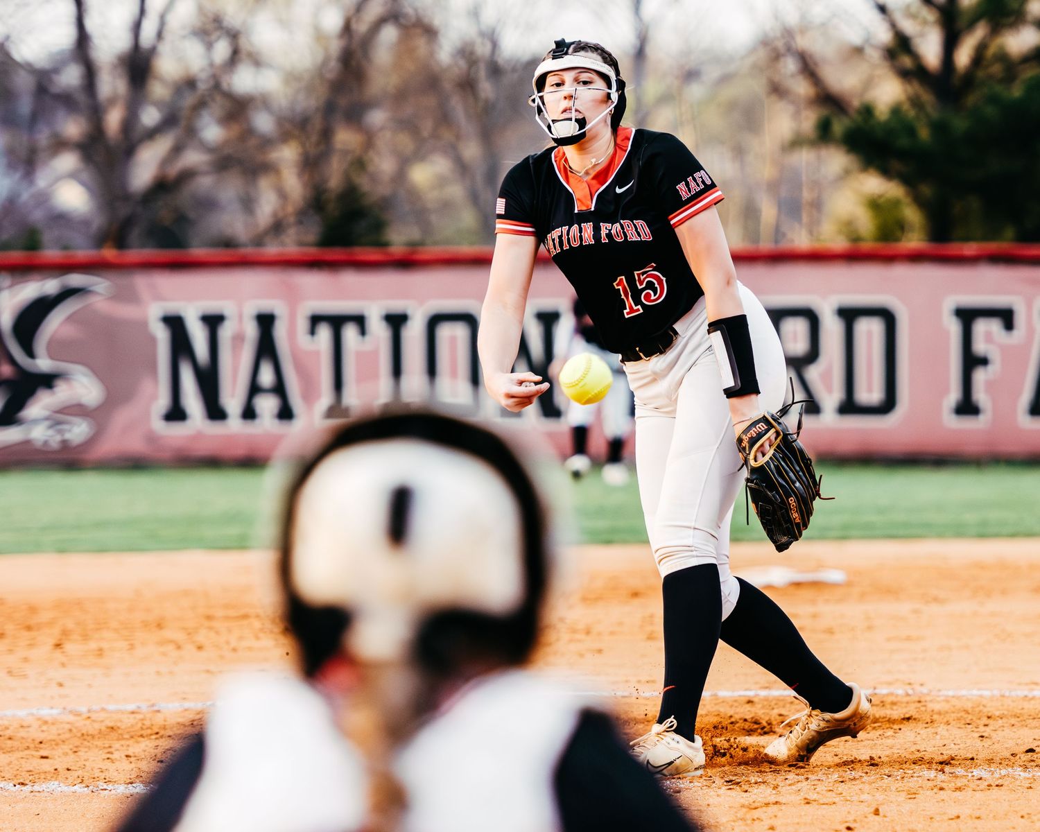 Nation Ford Falcons softball pitcher #15 delivers a pitch during a home game with the team banner visible in the background.