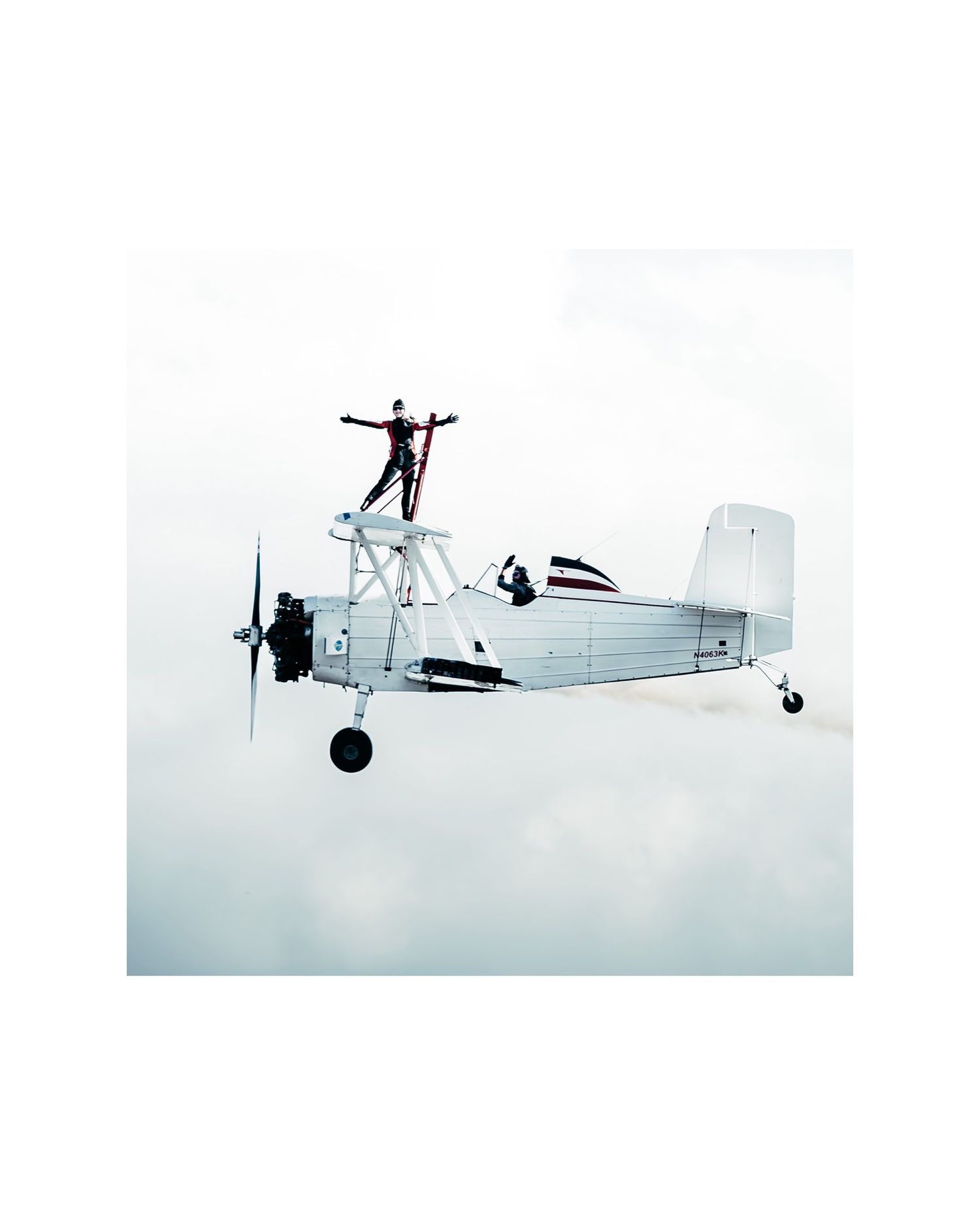 High-altitude shot of a wing walker standing on top of a white biplane soaring through a bright, cloudy sky.