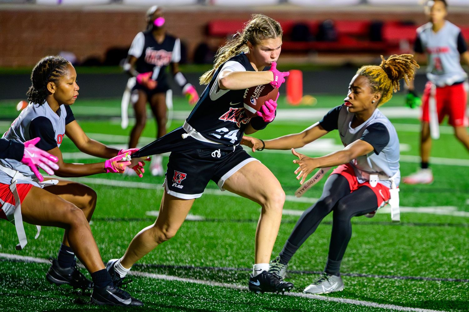 Girls' flag football action shot featuring a player in a black jersey number 24 weaving through defenders in grey and red.