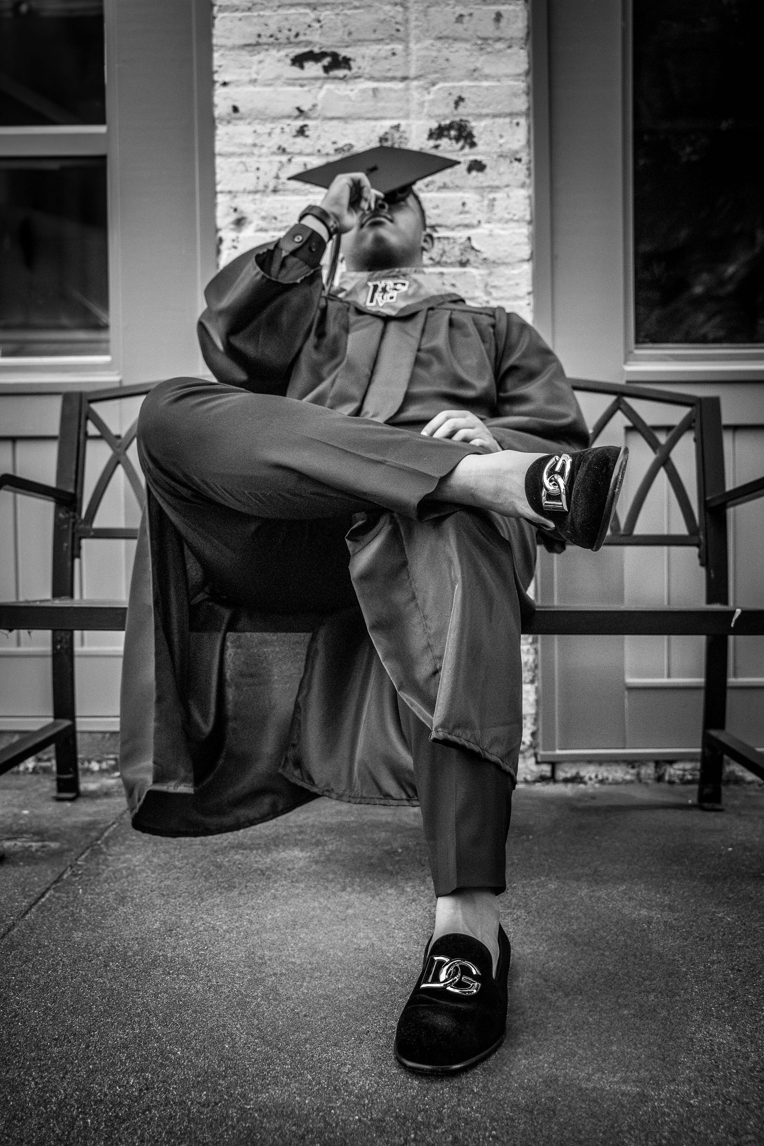 Black and white portrait of a graduate in a cap and gown sitting confidently on a bench with legs crossed.