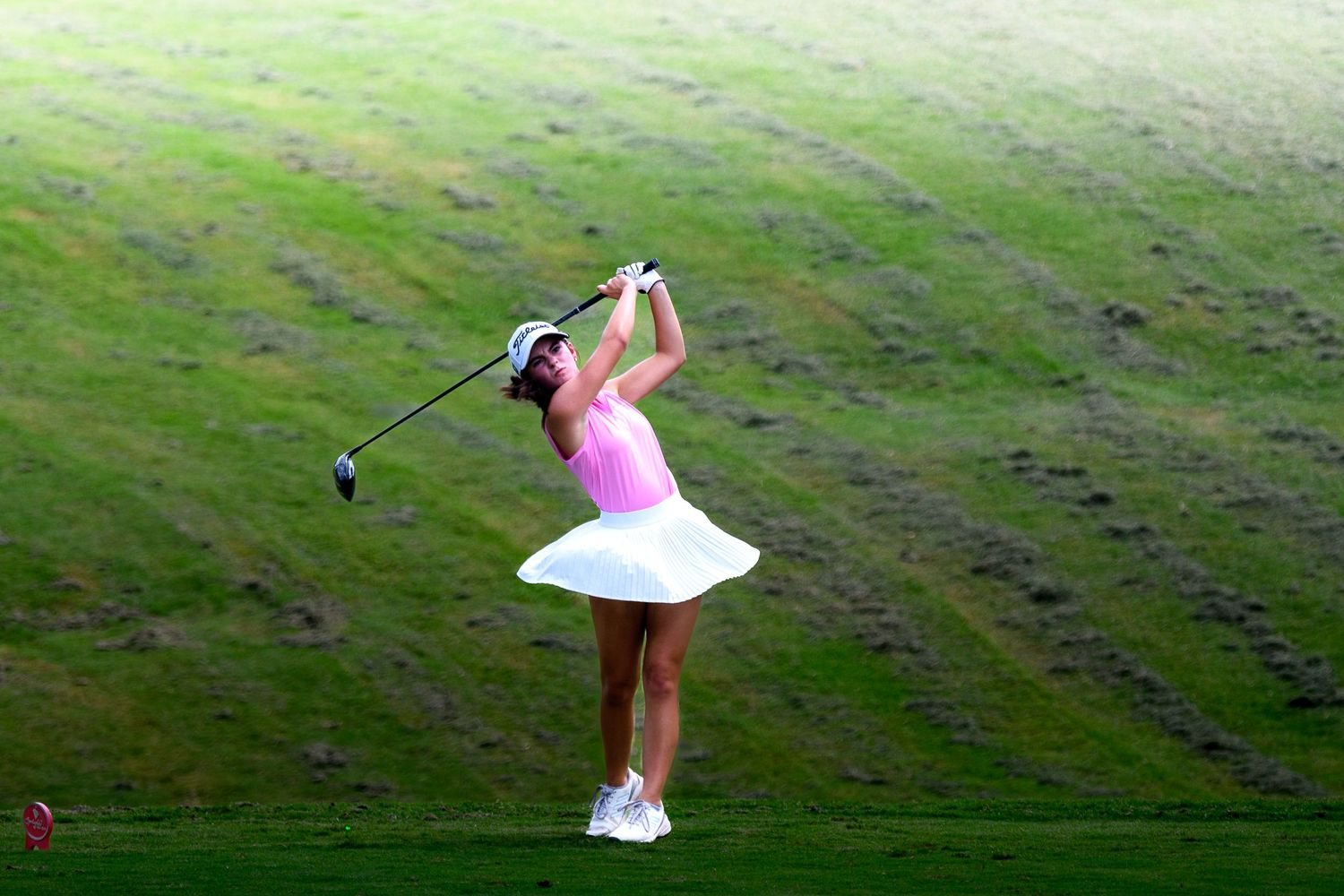 A female golfer in a pink top and white skirt completing a powerful drive on a lush green course.
