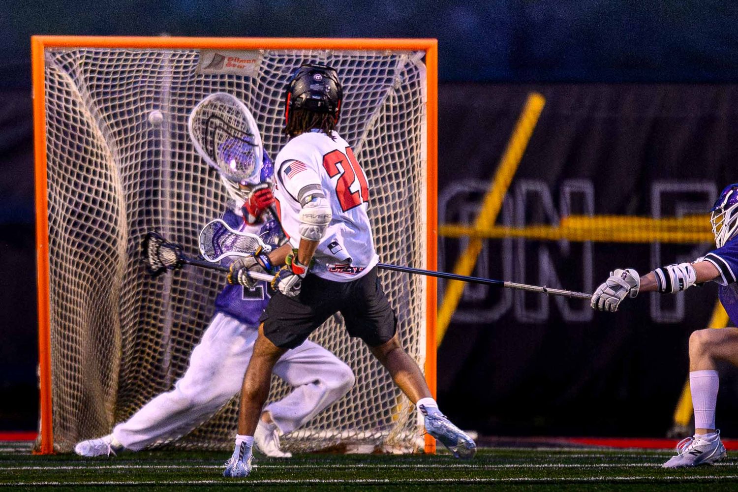 A lacrosse goalie in purple and white makes a dynamic save as an opponent in a white jersey looks on.