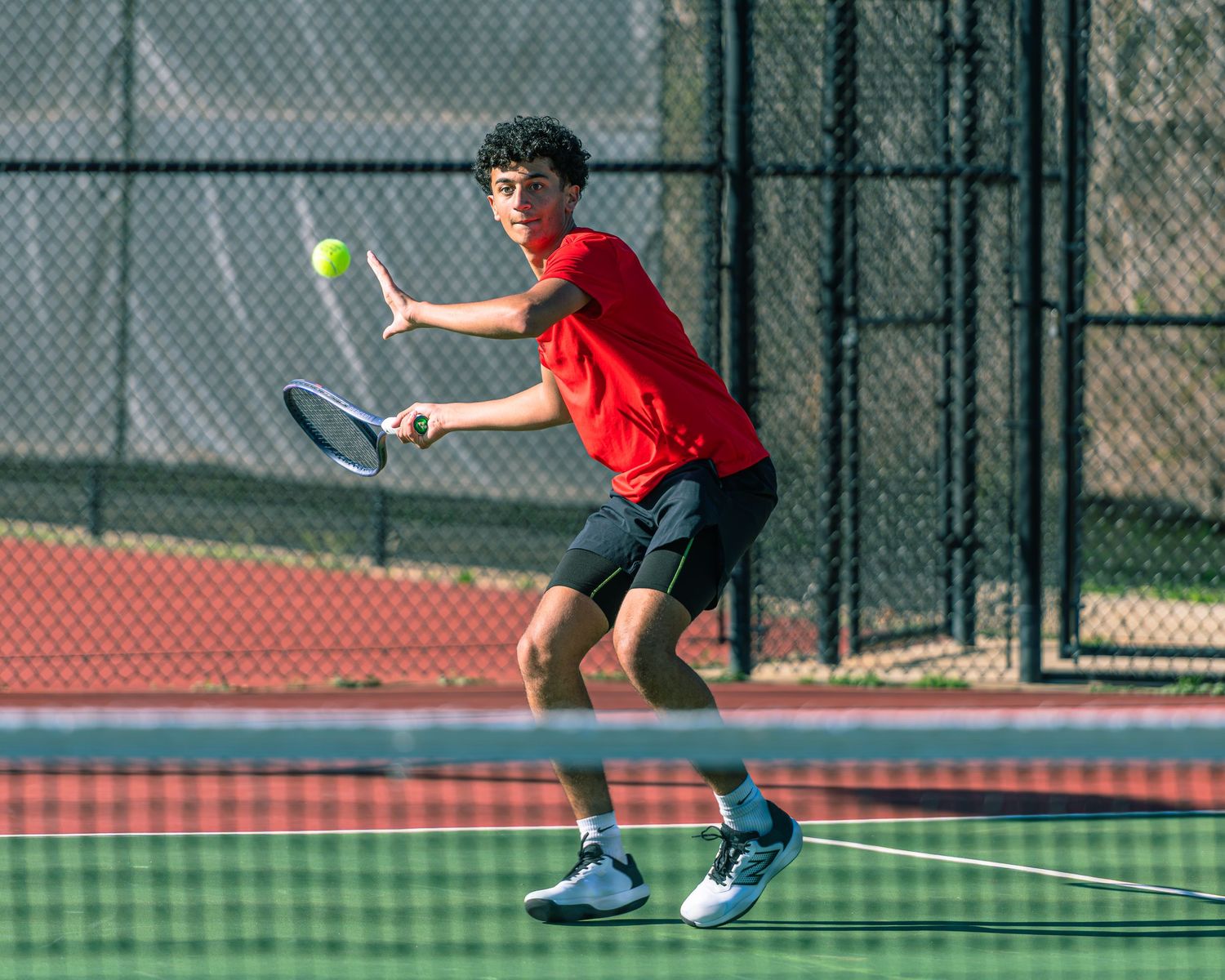 A high school boys tennis player in a red shirt prepares to hit a backhand shot during a match on an outdoor hard court.
