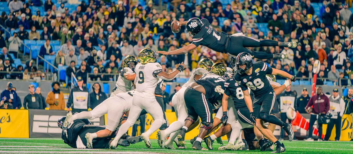 Dynamic action shot of a football player in a black jersey diving over a defensive line during a packed stadium game.
