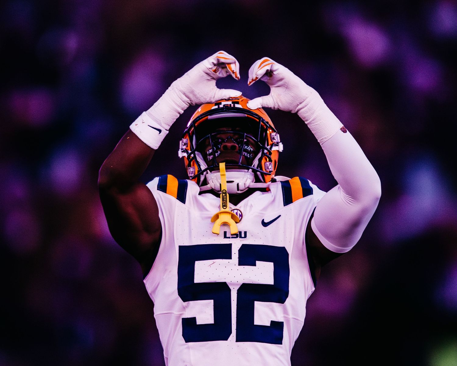 Close-up of an LSU football player in a white jersey number 52 making a heart gesture with his hands.