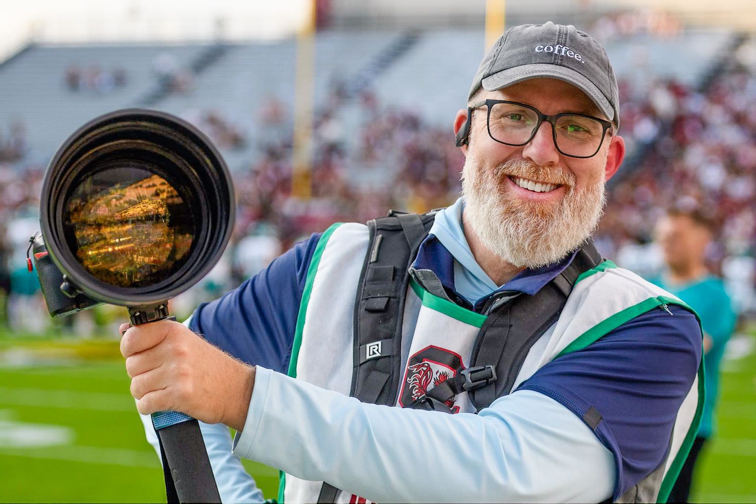 Professional sports photographer Chris Coffee smiles at a stadium while holding a Nikon camera with a 400mm telephoto lens on a monopod.