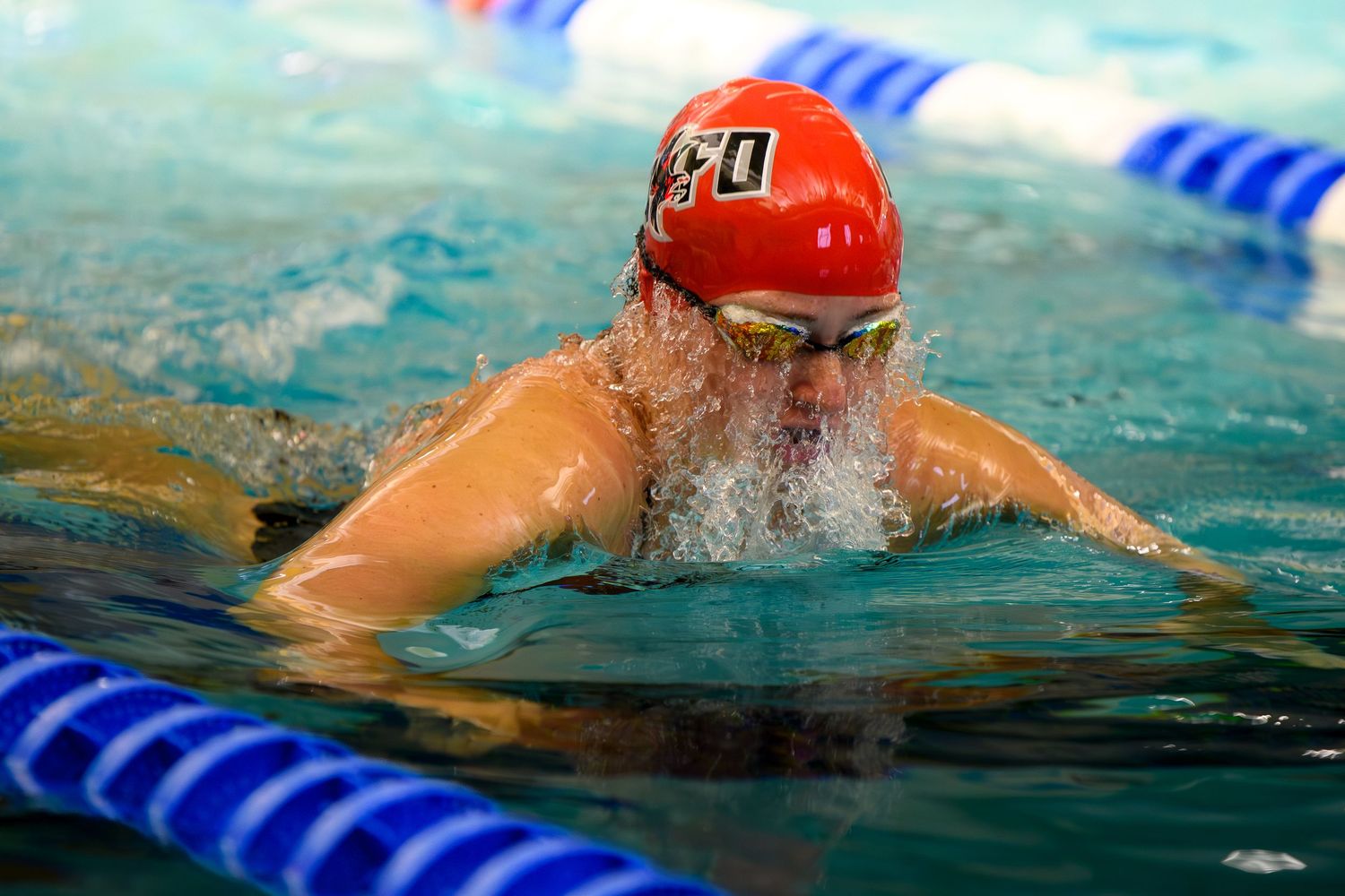 A swimmer in a red cap and goggles performing breaststroke, creating a splash of water at the surface.