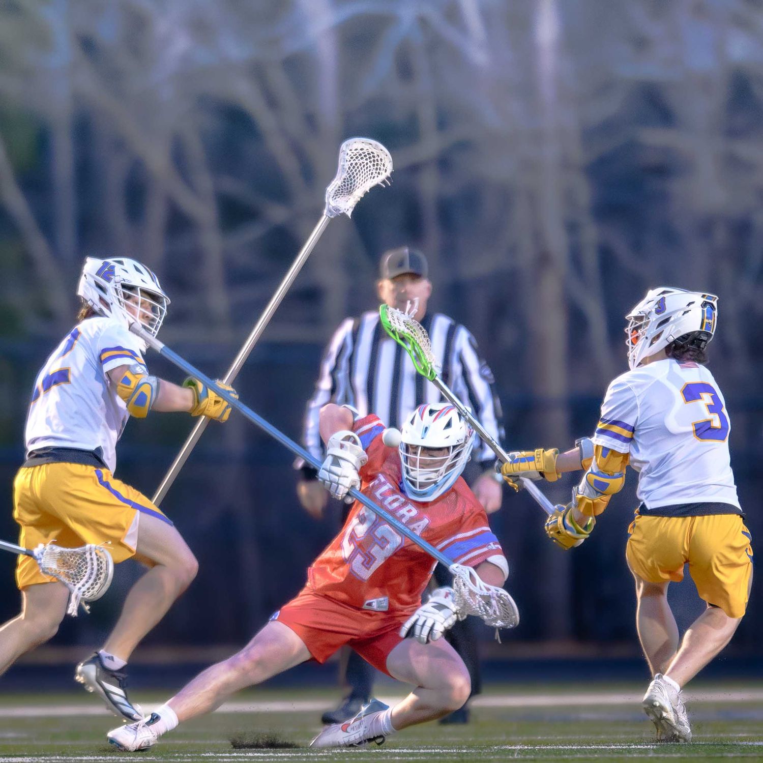 Intense lacrosse action showing three players competing for ball control with sticks raised during a game.