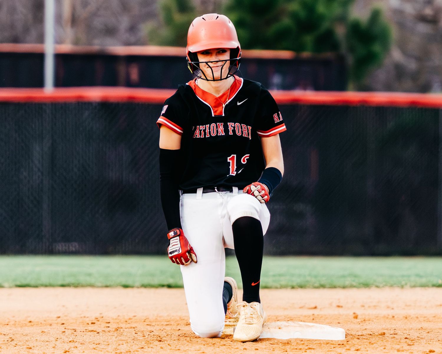 Nation Ford Falcons softball player #11 kneels on a base in her black and red jersey and orange helmet during a game.