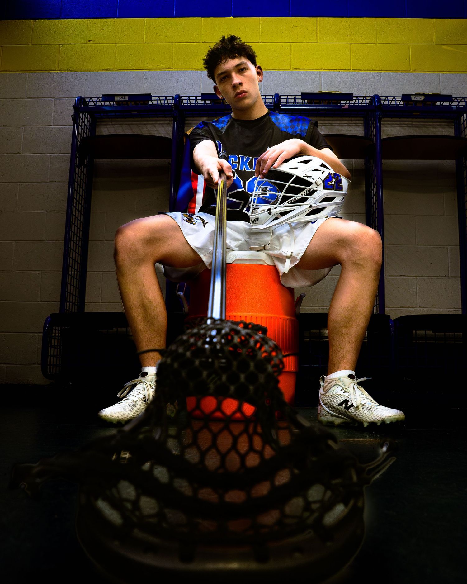 A lacrosse player sits in a locker room holding his helmet, with his stick positioned in the foreground.