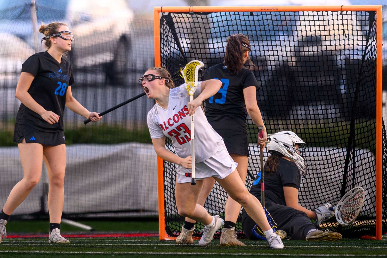 A female lacrosse player in a white jersey celebrating a goal in front of the net during a competitive match.