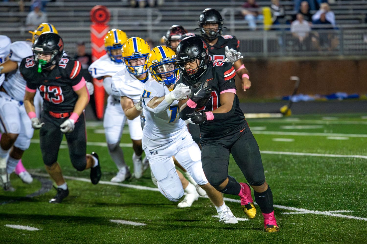 A football player in a black jersey with pink accents maneuvering past defenders in white jerseys on a turf field.