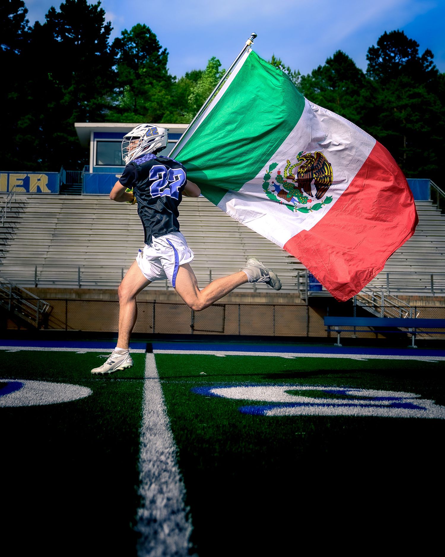 A lacrosse player in a white and blue uniform runs across the field carrying a large Mexican flag.