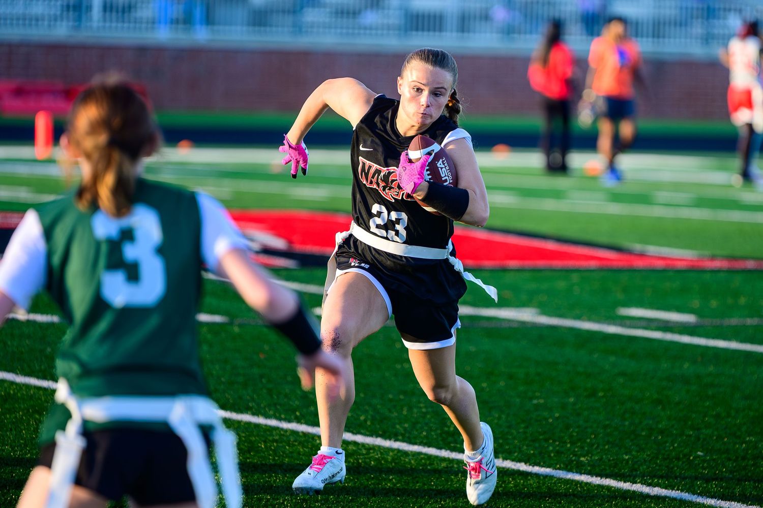 A girls' flag football player in a black jersey number 23 sprints downfield with the ball during a golden hour game.