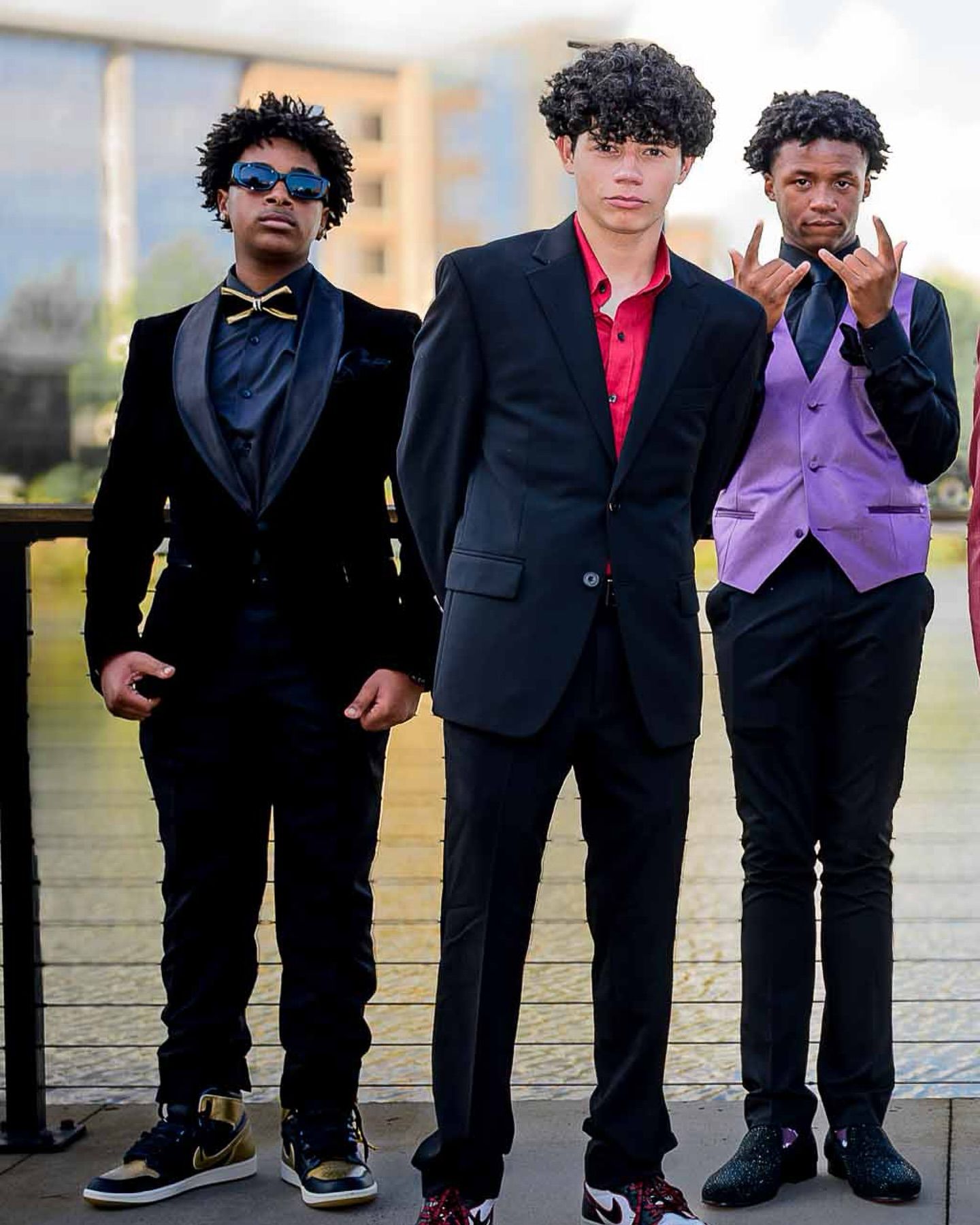 Three young men pose in formal attire: a black velvet tuxedo with gold accents, a black suit with a red shirt, and a purple vest with tie.