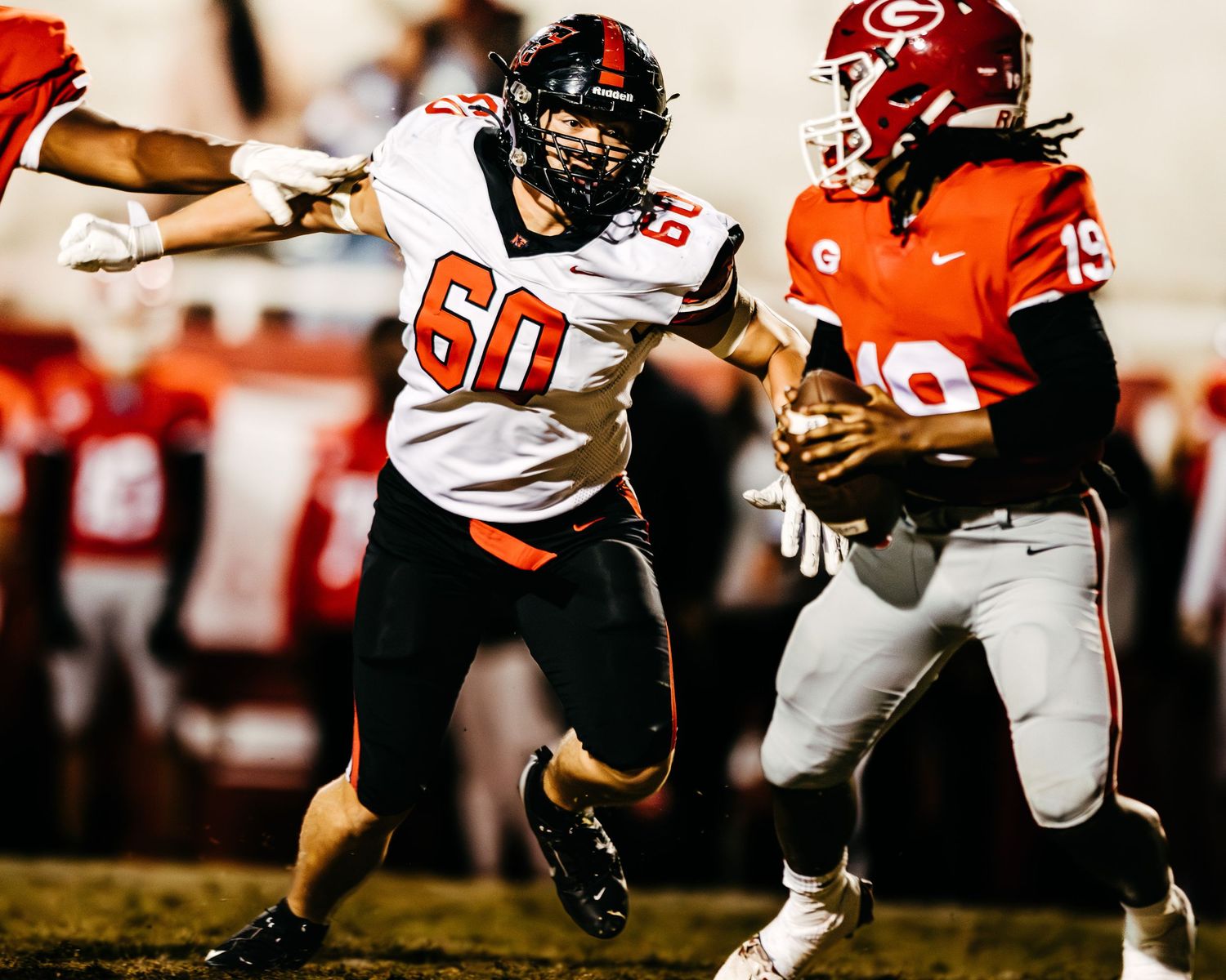A white-jerseyed football player number 60 blocks a red-jerseyed opponent during a night game under bright stadium lights.