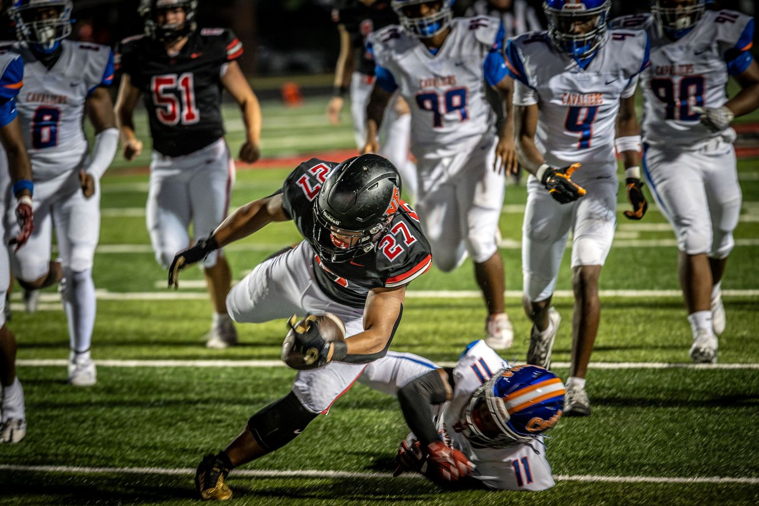 Action-packed high school football game under stadium lights with a running back in a black jersey diving past a defender for a touchdown.