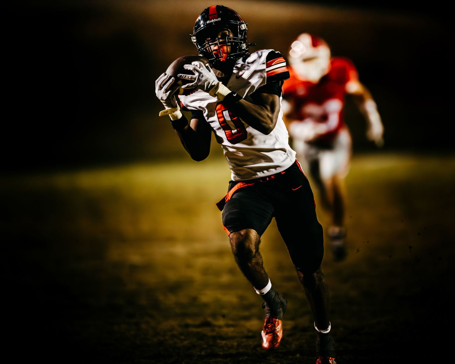 High school football player in a white jersey catching a pass during a night game under dramatic stadium lighting.