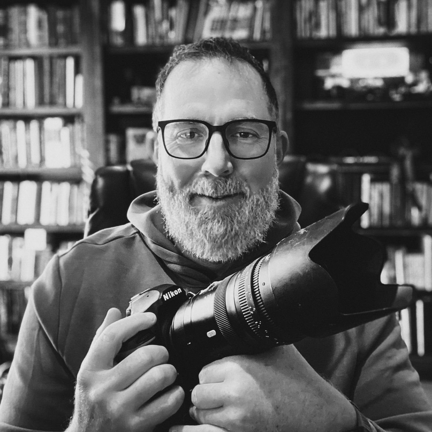 Black and white portrait of photographer Chris Coffee holding a Nikon camera with a large telephoto lens in front of a bookshelf.