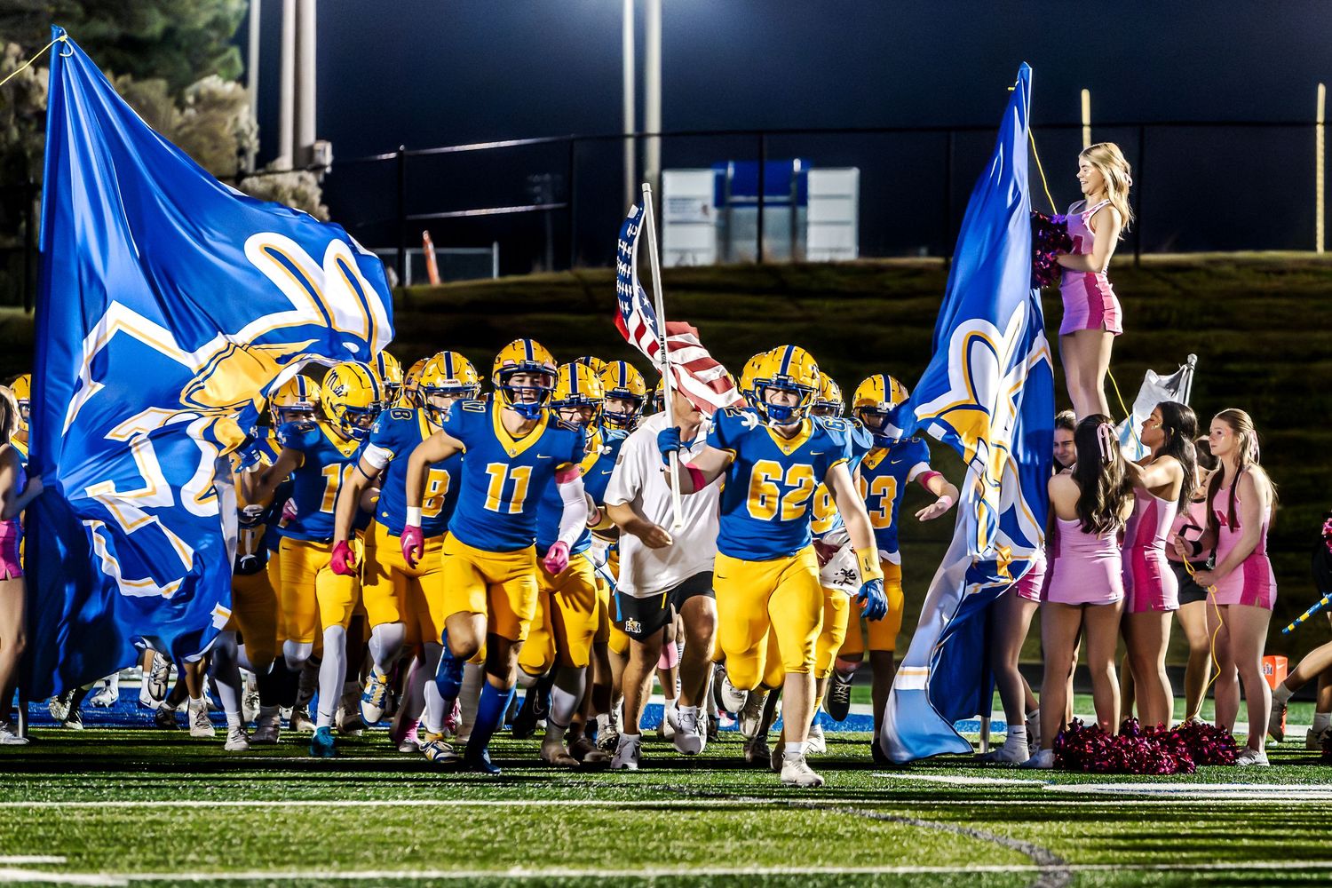 Fort Mill High School football players in blue and yellow uniforms sprint onto the field through large team flags.