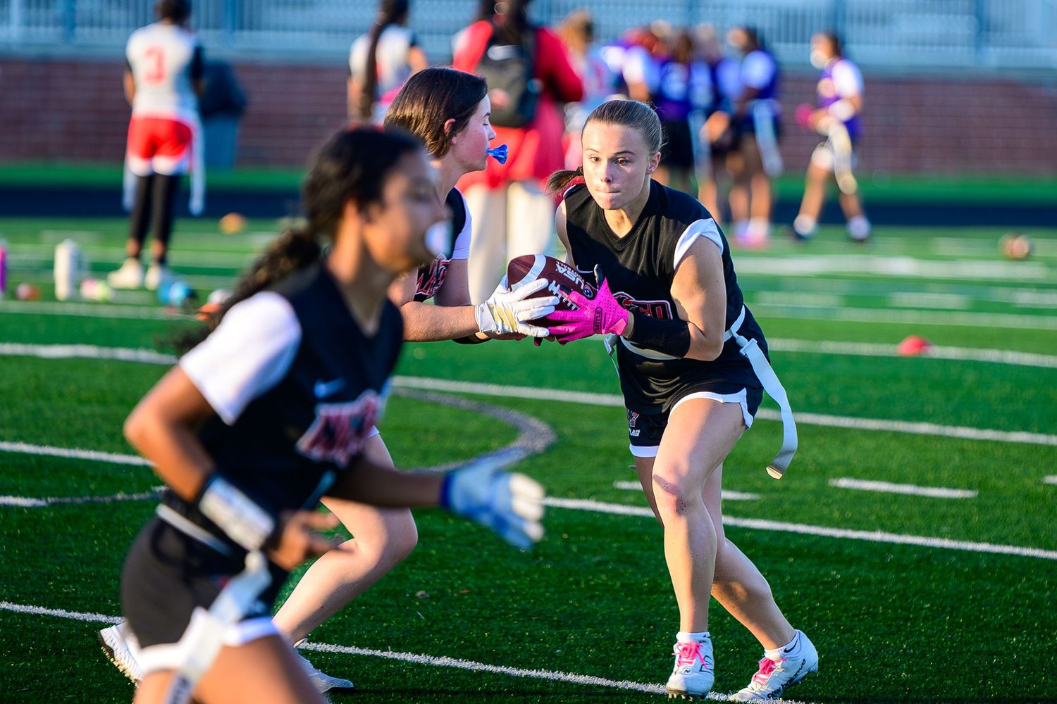 Female flag football player on the right receives a handoff from a teammate, wearing black jerseys and pink gloves on a green turf.