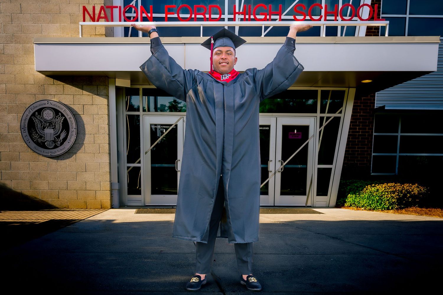 A graduate in a charcoal grey cap and gown poses with arms raised in front of the Nation Ford High School entrance.