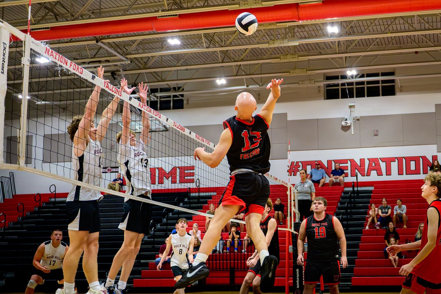 High school boys' volleyball player in a black jersey jumping for a spike against two blockers during a match in a gymnasium.