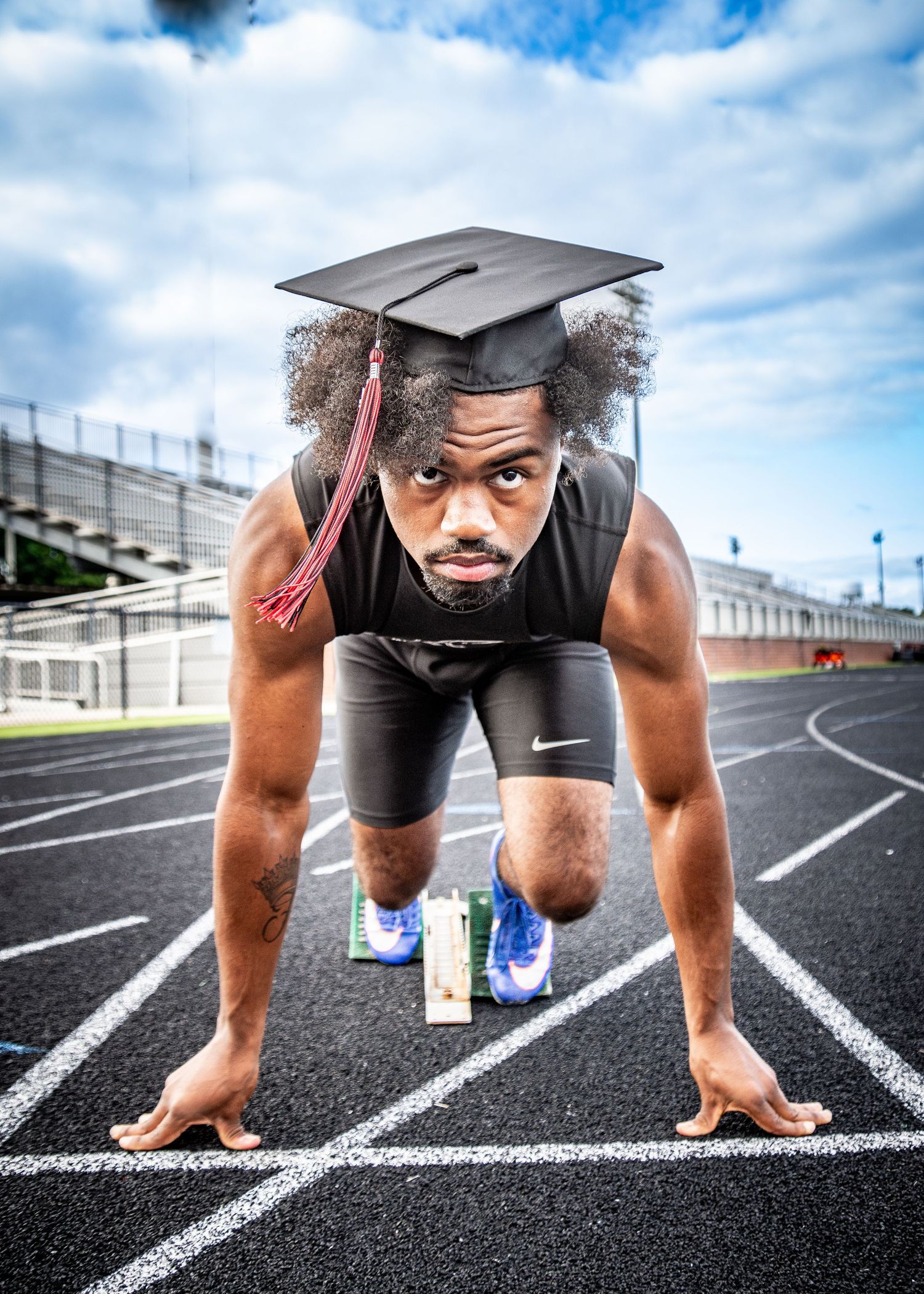 A young Black graduate in a track starting position on a stadium lane, wearing a graduation cap and athletic gear.
