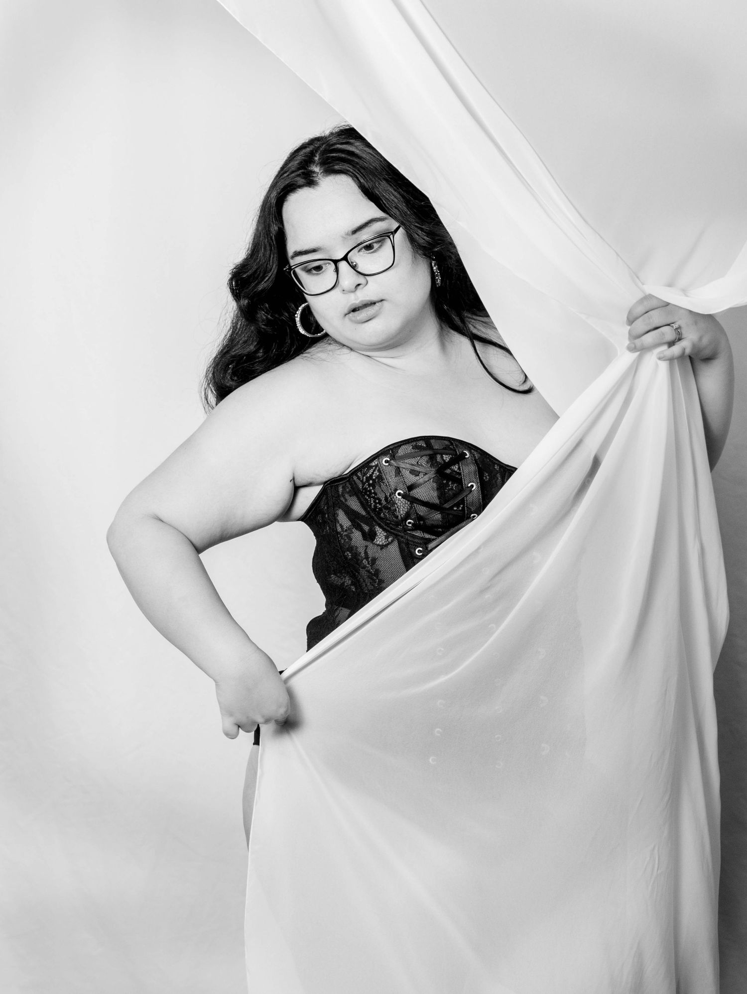 Black and white boudoir portrait of a woman in glasses and a black lace corset, artfully holding sheer white fabric in an elegant studio set