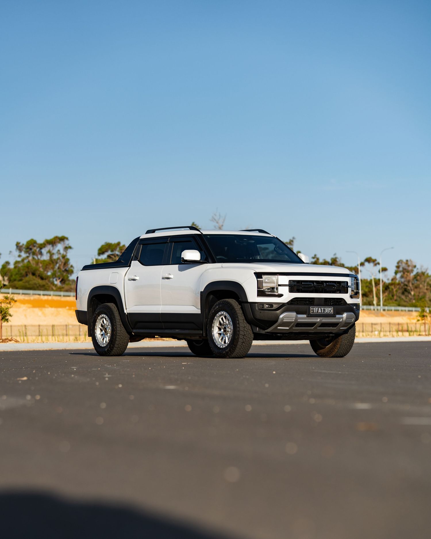White pickup truck parked on pavement under clear blue sky, showcasing rugged off-road design with roof rack and bold front fascia.