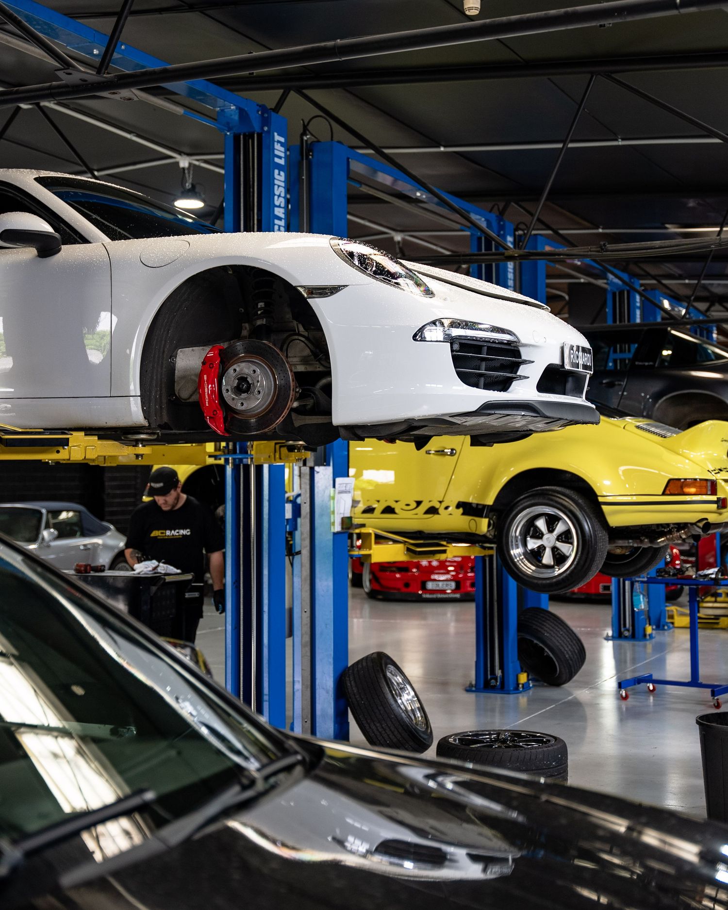 Busy auto repair shop with white Porsche SUV on blue lift, yellow classic car raised, and mechanics working on vehicles.
