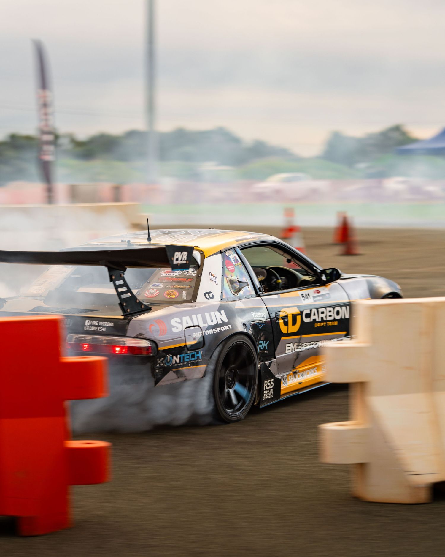 A drift car with Carbon sponsorship slides around a track corner, producing tire smoke at a motorsport competition.