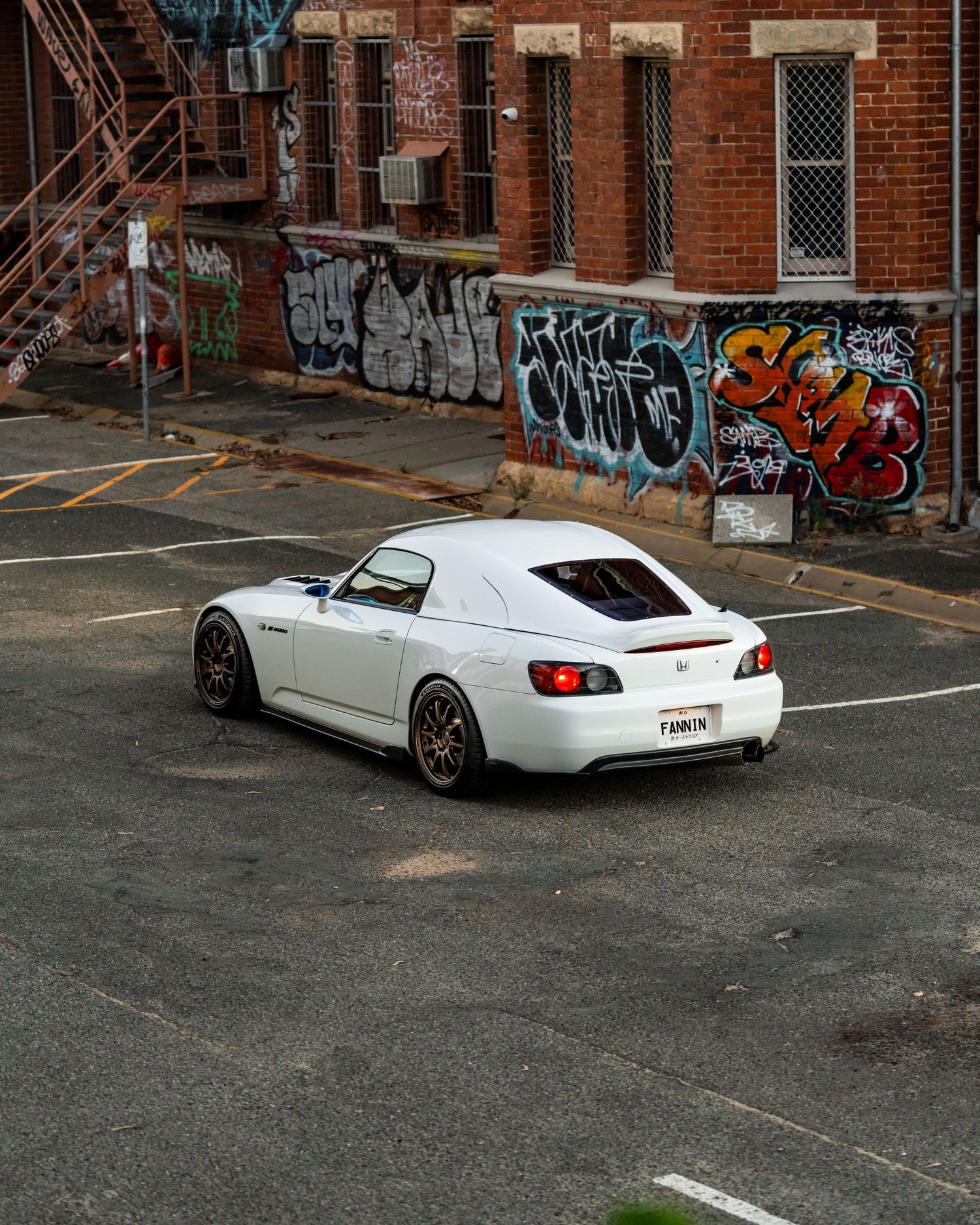 White Honda S2000 with bronze wheels parked in urban lot with colorful graffiti wall backdrop.