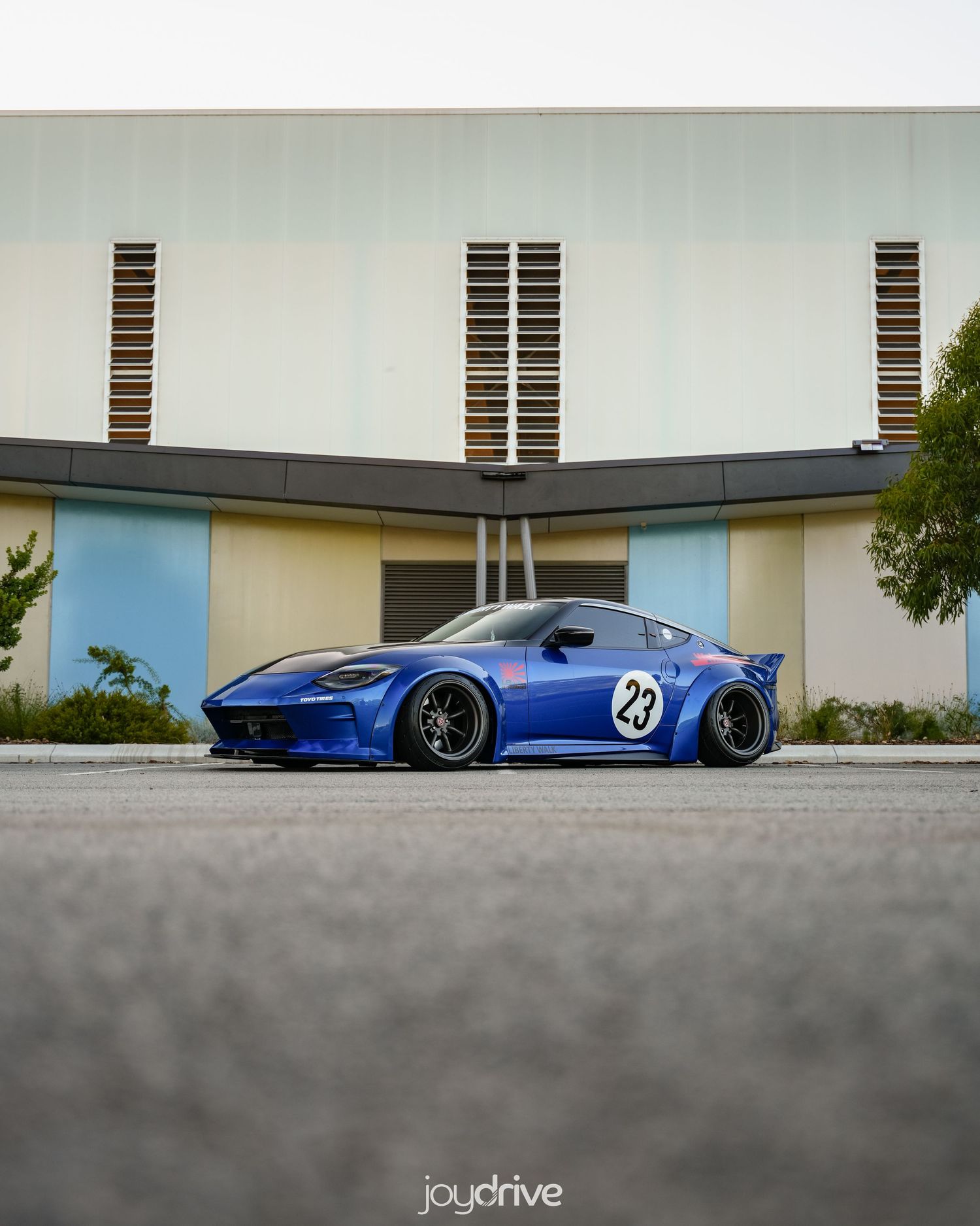 Modified blue Nissan Z with wide body kit and number 23 parked outside a modern building.