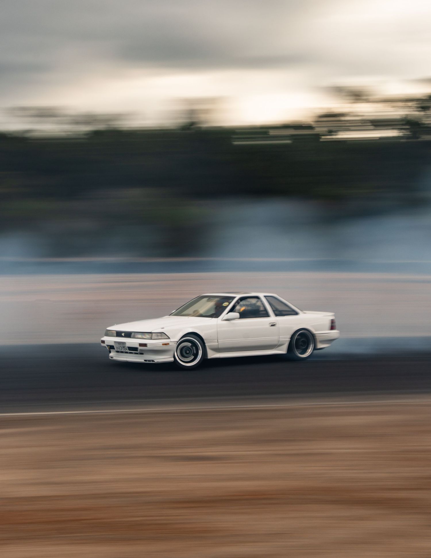 White Toyota Soarer drifting on a dirt track at dusk with smoke.