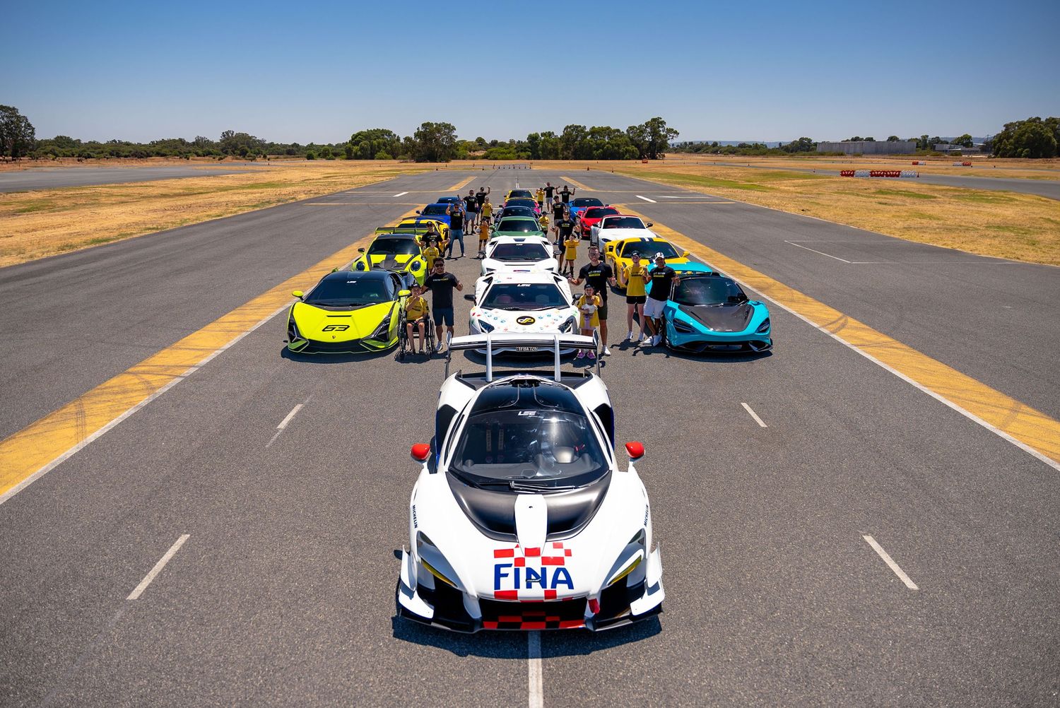 Lineup of colorful supercars on a runway with drivers standing beside them on a sunny day.