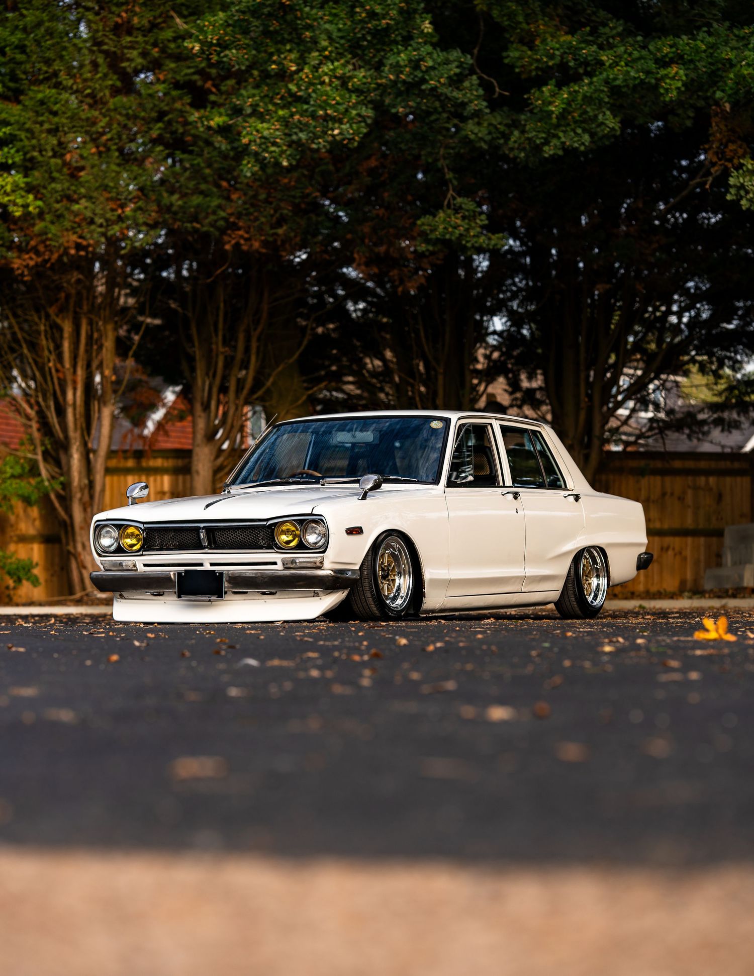 Classic white Nissan Skyline slammed low with gold wheels parked on a quiet street.