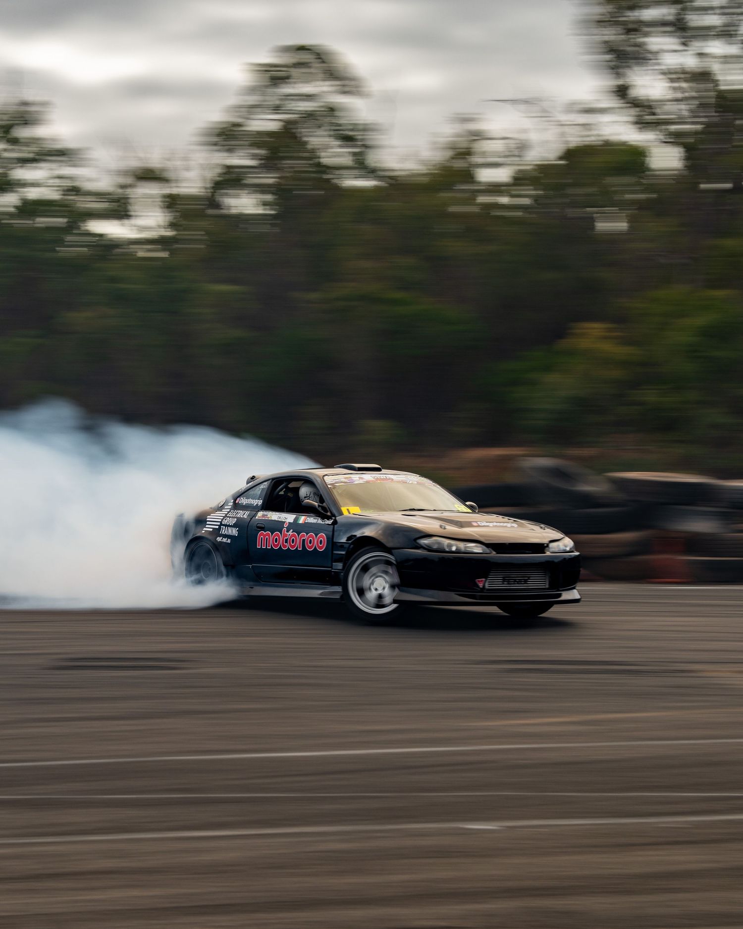 Black drift car producing thick white smoke while drifting on a track under cloudy skies.