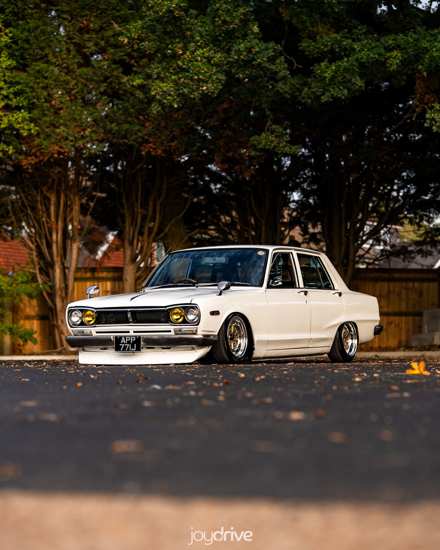 Slammed white classic Nissan Bluebird 510 with gold wheels parked on a suburban street.