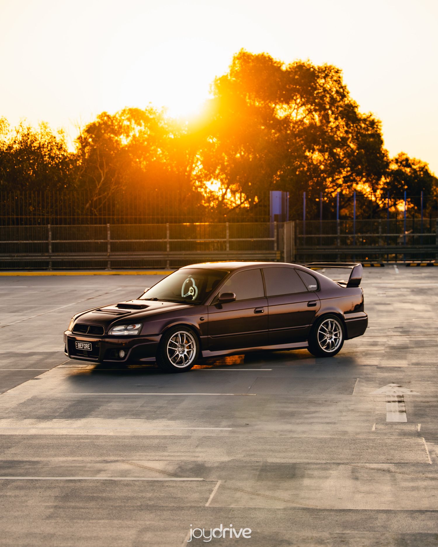 Dark purple Subaru Liberty sedan parked on rooftop with golden sunset glowing through trees.