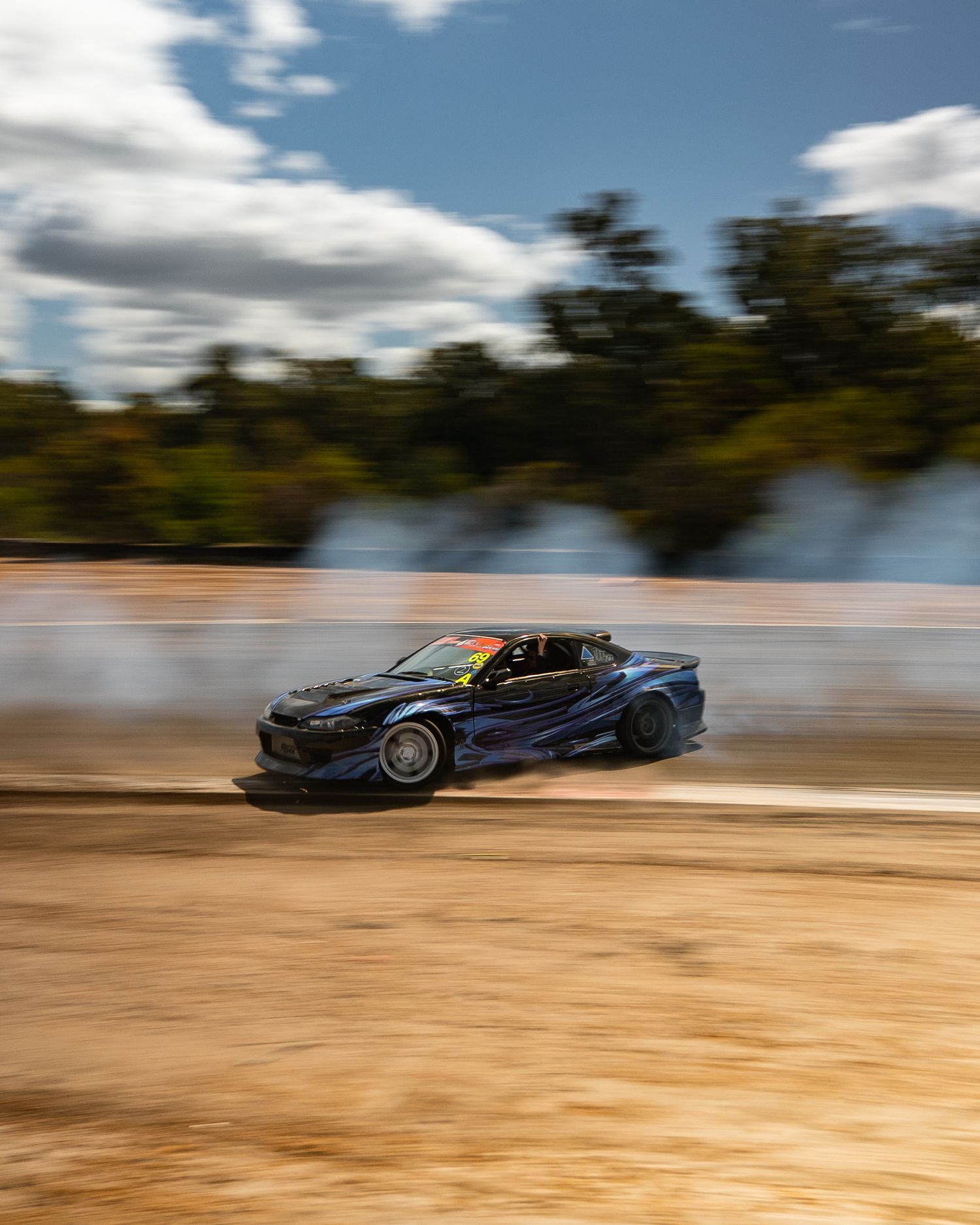 Blue drift car sliding sideways on a dirt track producing large smoke clouds.