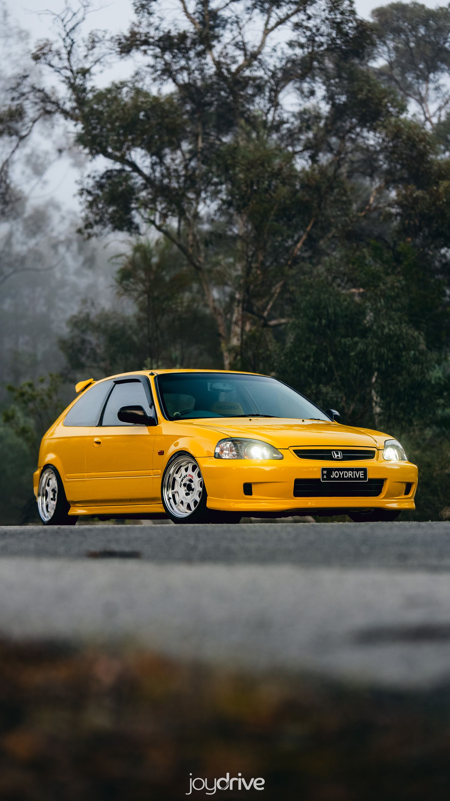 Bright yellow modified Honda Civic EK hatchback on white wheels parked on a misty road.