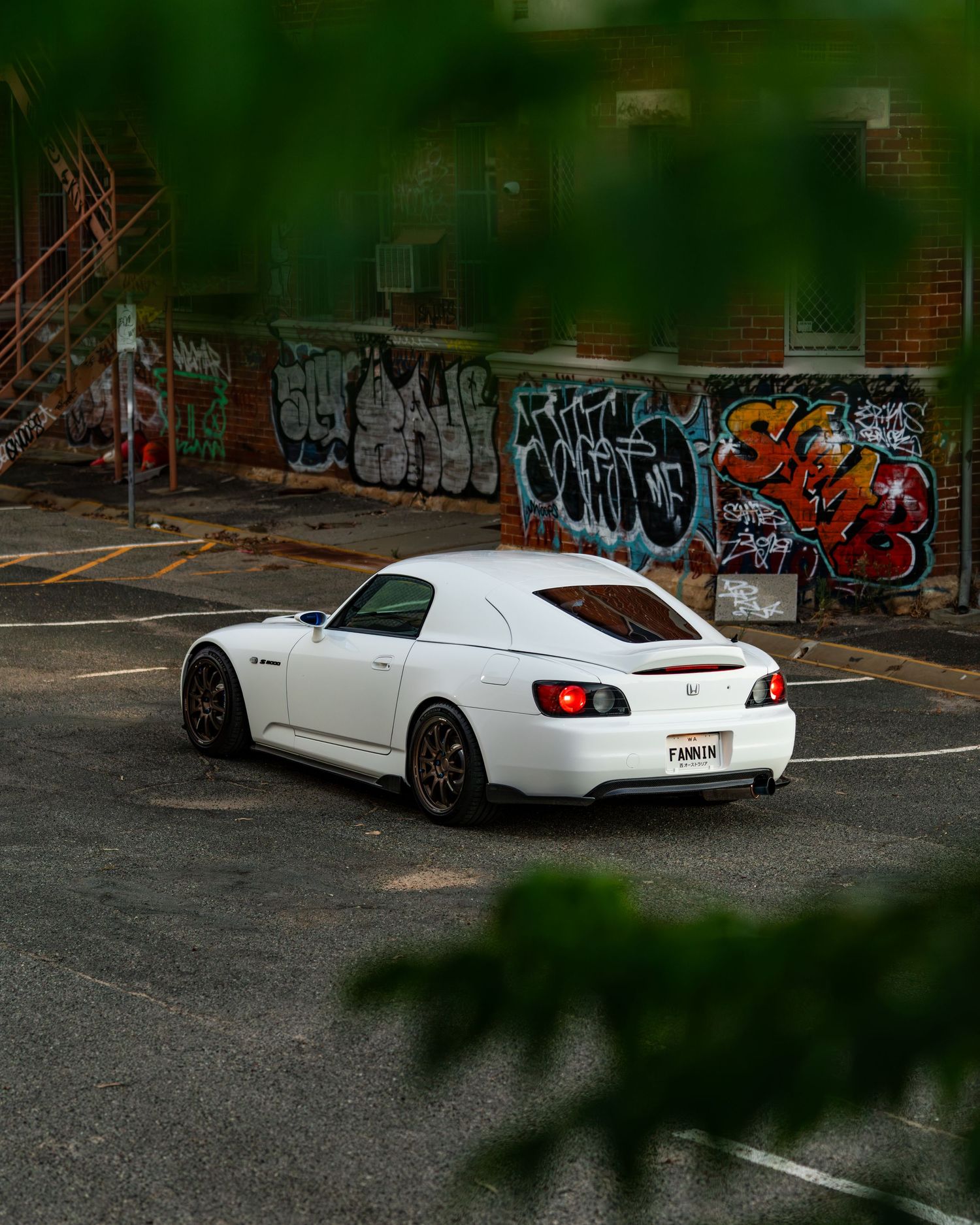 White Honda S2000 parked in a lot with colorful graffiti-covered brick wall in background.