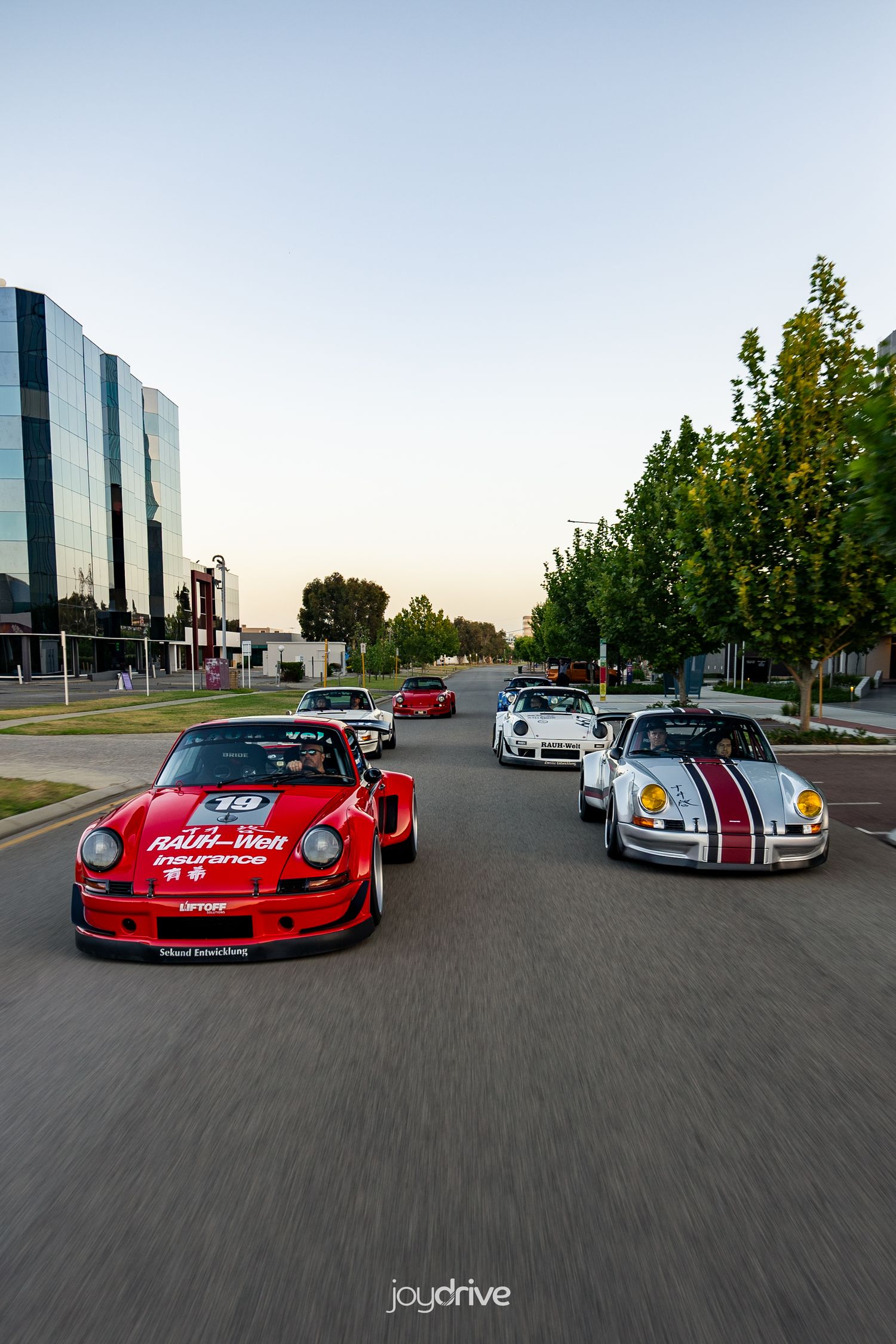 Group of classic Porsche 911 RWB cars driving through urban street at dusk.
