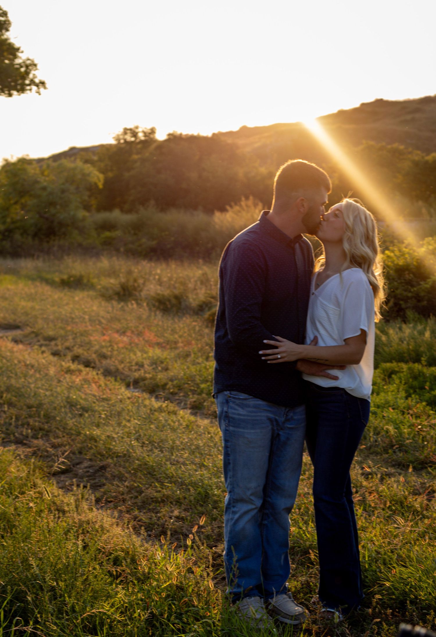 An engaged couple shares a romantic kiss in a sunlit field with golden sunset rays behind them.
