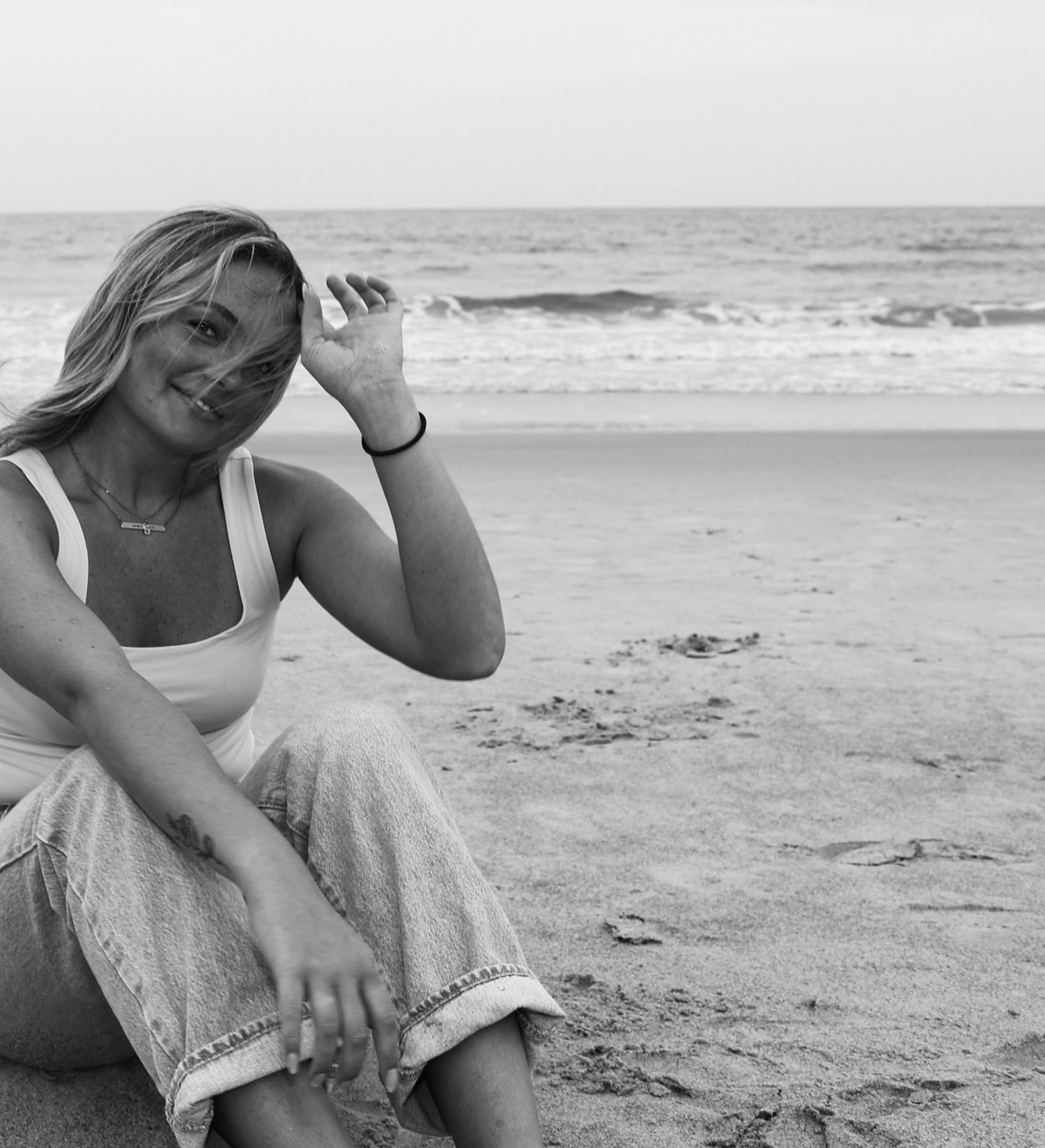 Black and white portrait of smiling blonde woman sitting on sandy beach with ocean waves behind her.