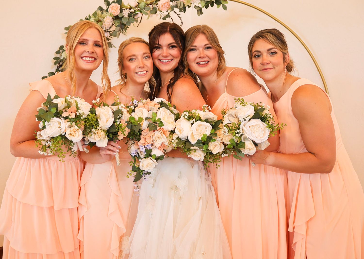 A bride poses with four bridesmaids in peach dresses holding white floral bouquets under a gold arch.